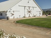 a white barn in a large field surrounded by flowers and greenery at the end of a driveway