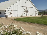 a white barn in a large field surrounded by flowers and greenery at the end of a driveway