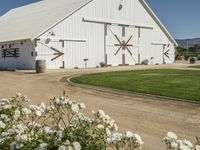 a white barn in a large field surrounded by flowers and greenery at the end of a driveway