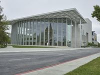 the building is made of glass with trees growing on both sides of the sidewalk and road side