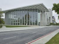 the building is made of glass with trees growing on both sides of the sidewalk and road side