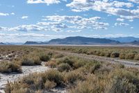 the road winds through a high desert landscape, under a cloudy blue sky with clouds