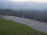 the motorcycle is on the road by the grassy hill side and a bench overlooking the view