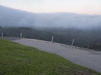 the motorcycle is on the road by the grassy hill side and a bench overlooking the view