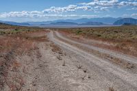 dirt road in an arid field with mountains in background at daytime with cloudy blue skies