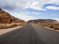 a lone highway in the middle of desert land with cliffs in the background, and clouds above