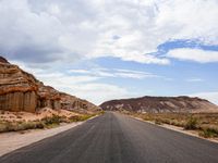 a lone highway in the middle of desert land with cliffs in the background, and clouds above