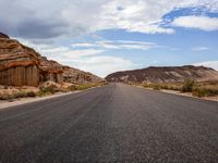 a lone highway in the middle of desert land with cliffs in the background, and clouds above