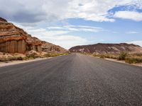 a lone highway in the middle of desert land with cliffs in the background, and clouds above