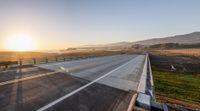 the sun shining on an empty highway at sunset with hills in background and fenced field in foreground