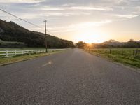 an empty paved road in front of a fence and mountains at sunset, with clouds overhead