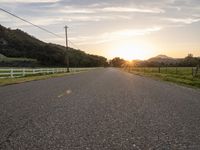 an empty paved road in front of a fence and mountains at sunset, with clouds overhead