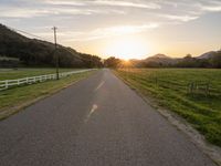 an empty paved road in front of a fence and mountains at sunset, with clouds overhead