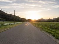 an empty paved road in front of a fence and mountains at sunset, with clouds overhead