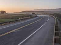 an empty highway surrounded by hills and grass at sunsettime by a fenced area