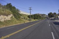 a long empty road passing along side of a hill with power lines on it and grass