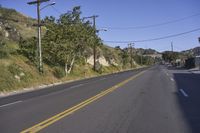 a long empty road passing along side of a hill with power lines on it and grass