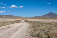 a dirt road running through a dry grassy field with a blue sky and mountains in the background