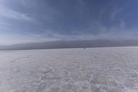 two people stand on a salt covered desert plain under blue skies and clouds, with mountains in the distance