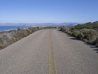 a lone road lined with bushes by the ocean next to some mountains that have mountains on one side