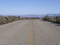 a lone road lined with bushes by the ocean next to some mountains that have mountains on one side