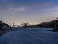 the moon rising over a valley and a road at dusk, with trees and fence surrounding it