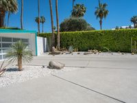 a planter and rocks are in the driveway by a green hedge and a white stucco house