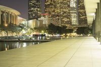 a long sidewalk near a body of water with tall buildings behind it at night time