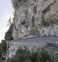 the person is riding their motorcycle on the side of a road in the woods with a view of mountains and cliff