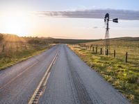 a long road leads to a windmill in the distance, and grass and a fence are growing at the other end