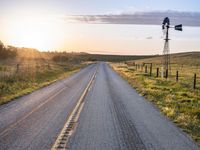 a long road leads to a windmill in the distance, and grass and a fence are growing at the other end