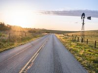 a long road leads to a windmill in the distance, and grass and a fence are growing at the other end