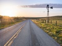 a long road leads to a windmill in the distance, and grass and a fence are growing at the other end