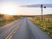 a long road leads to a windmill in the distance, and grass and a fence are growing at the other end