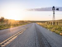 a long road leads to a windmill in the distance, and grass and a fence are growing at the other end