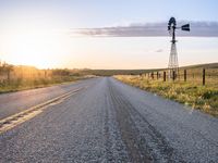 a long road leads to a windmill in the distance, and grass and a fence are growing at the other end