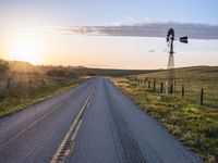 a long road leads to a windmill in the distance, and grass and a fence are growing at the other end