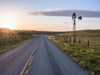 a long road leads to a windmill in the distance, and grass and a fence are growing at the other end