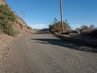 a narrow dirt road is surrounded by shrubs and power lines above the hill top with telephone wires on each side