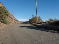 a narrow dirt road is surrounded by shrubs and power lines above the hill top with telephone wires on each side