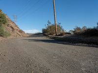 a narrow dirt road is surrounded by shrubs and power lines above the hill top with telephone wires on each side