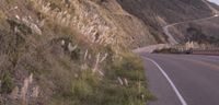 a mountain road on a hillside with tall grass along side of road and fenced area