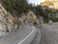 the road is curving past a steep cliff side and green pine trees grow on both sides