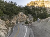 the road is curving past a steep cliff side and green pine trees grow on both sides