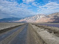 dirt road with mountains in background under blue sky and clouds over water with dirt road in foreground