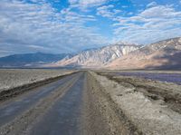 dirt road with mountains in background under blue sky and clouds over water with dirt road in foreground