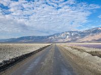 dirt road with mountains in background under blue sky and clouds over water with dirt road in foreground