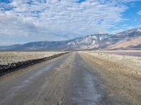 dirt road with mountains in background under blue sky and clouds over water with dirt road in foreground