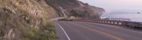 a curved road on the side of a mountain overlooking a ocean cliff, in motion with the car going on