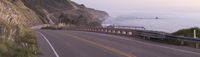 a curved road on the side of a mountain overlooking a ocean cliff, in motion with the car going on